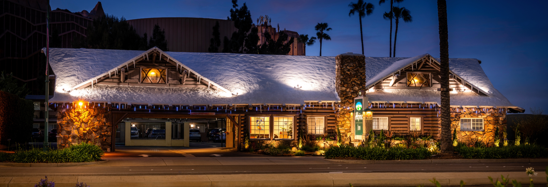 A log cabin-style building with snow on the roof and lights on at dusk.