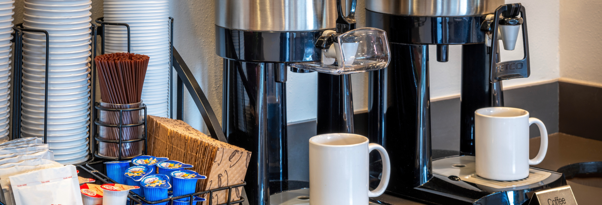 A coffee station with two coffee makers, cups, sugar packets, and creamers.