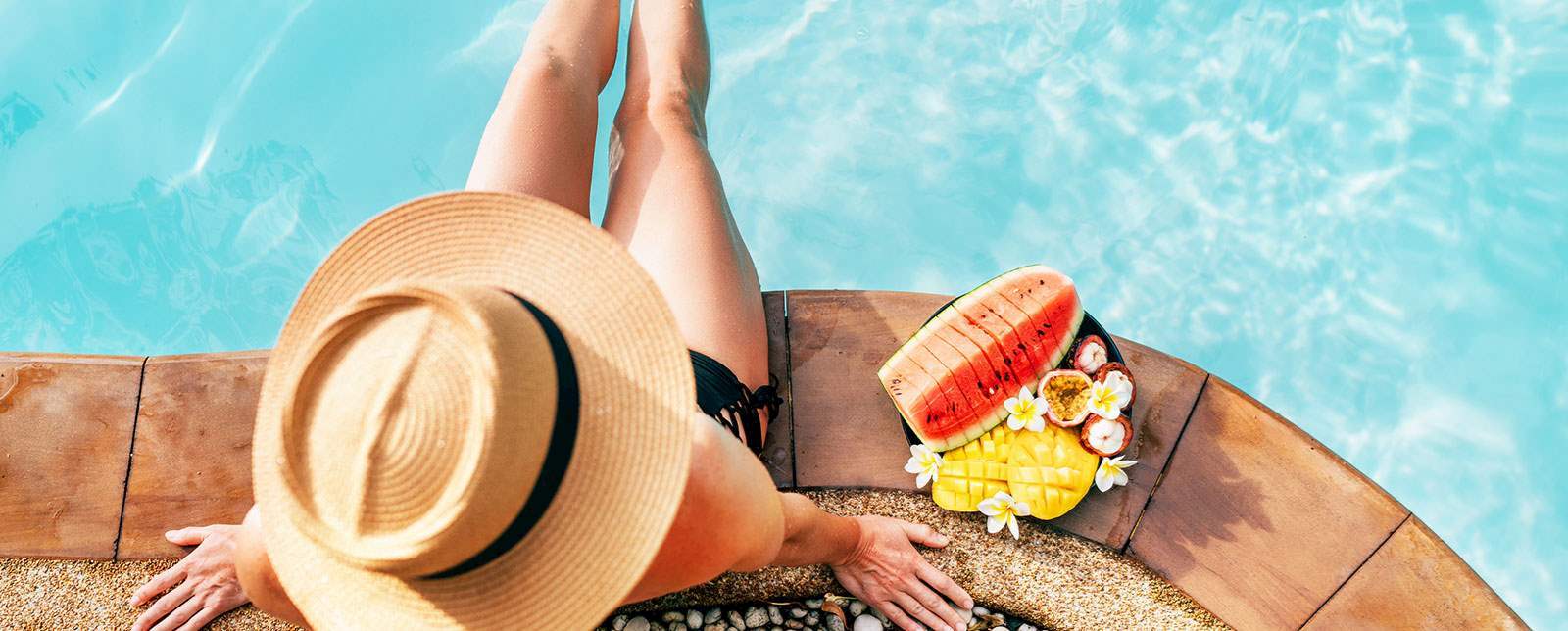 A woman in a hat is sitting on the edge of a pool with fruits.